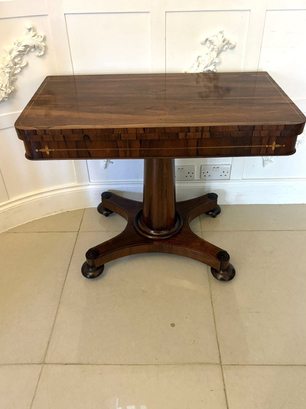 A Regency rosewood card table featuring fine brass inlay stringing, a fold-over swivel top, a central pedestal stem, and a quadform base on round feet. View XI
