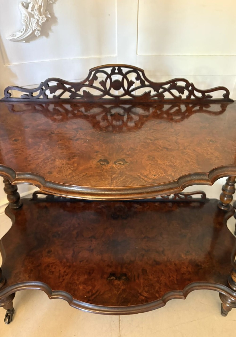 A pair of Victorian three-tier burr walnut whatnots featuring serpentine shelves, fretwork crestings, and turned support columns on castors. View V