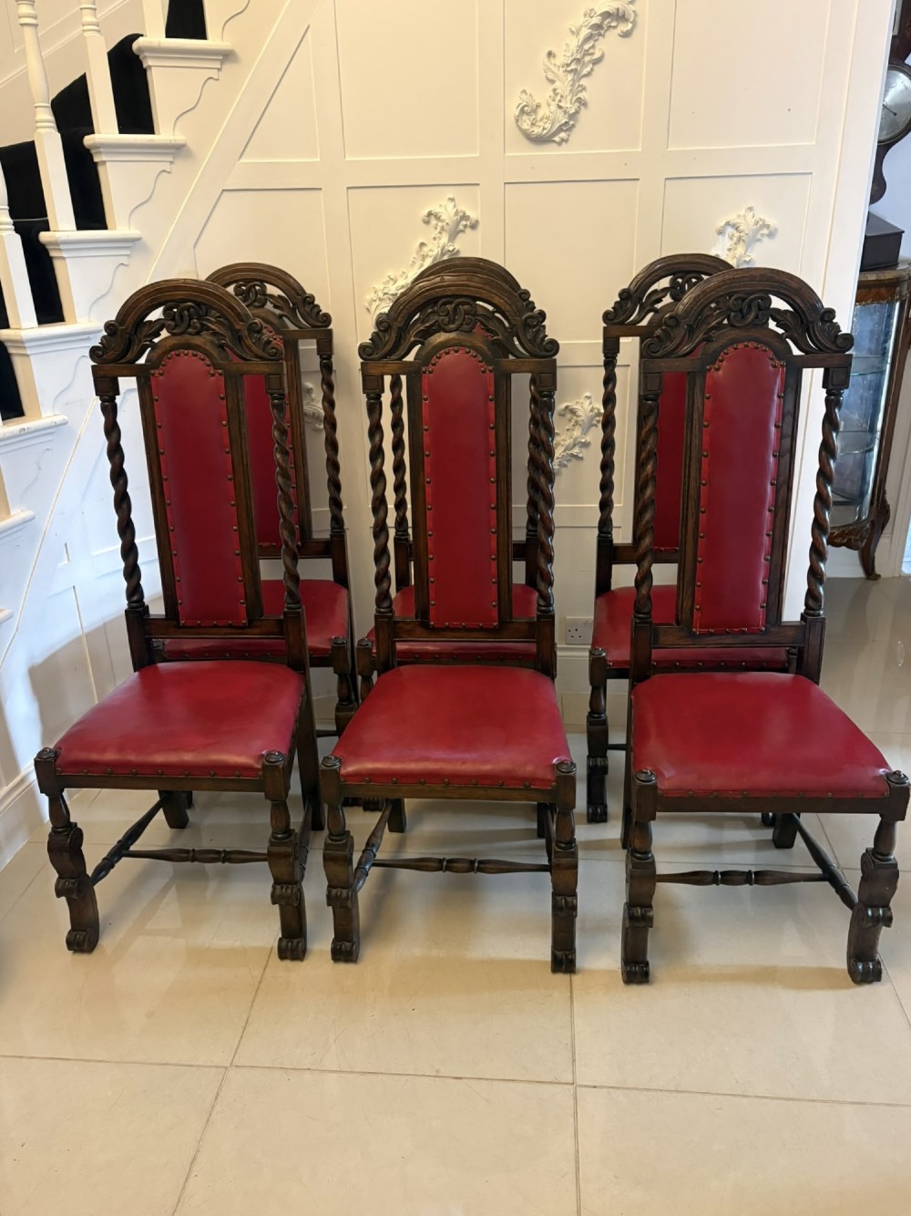 A set of high-backed dark oak dining chairs with barley twist uprights, carved cresting rails, and red upholstered seats and back panels with stud trim. View VII
