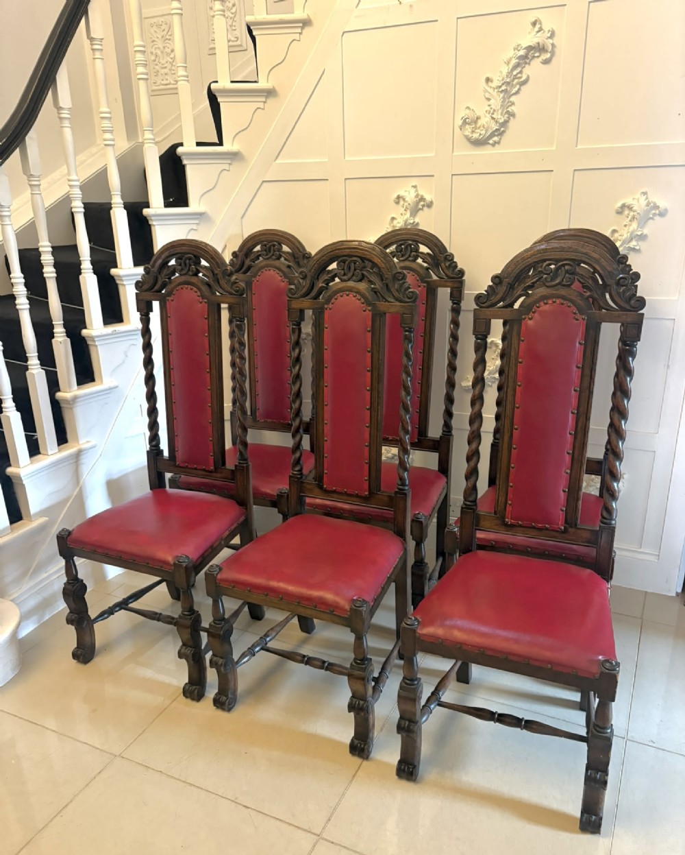 A set of high-backed dark oak dining chairs with barley twist uprights, carved cresting rails, and red upholstered seats and back panels with stud trim.