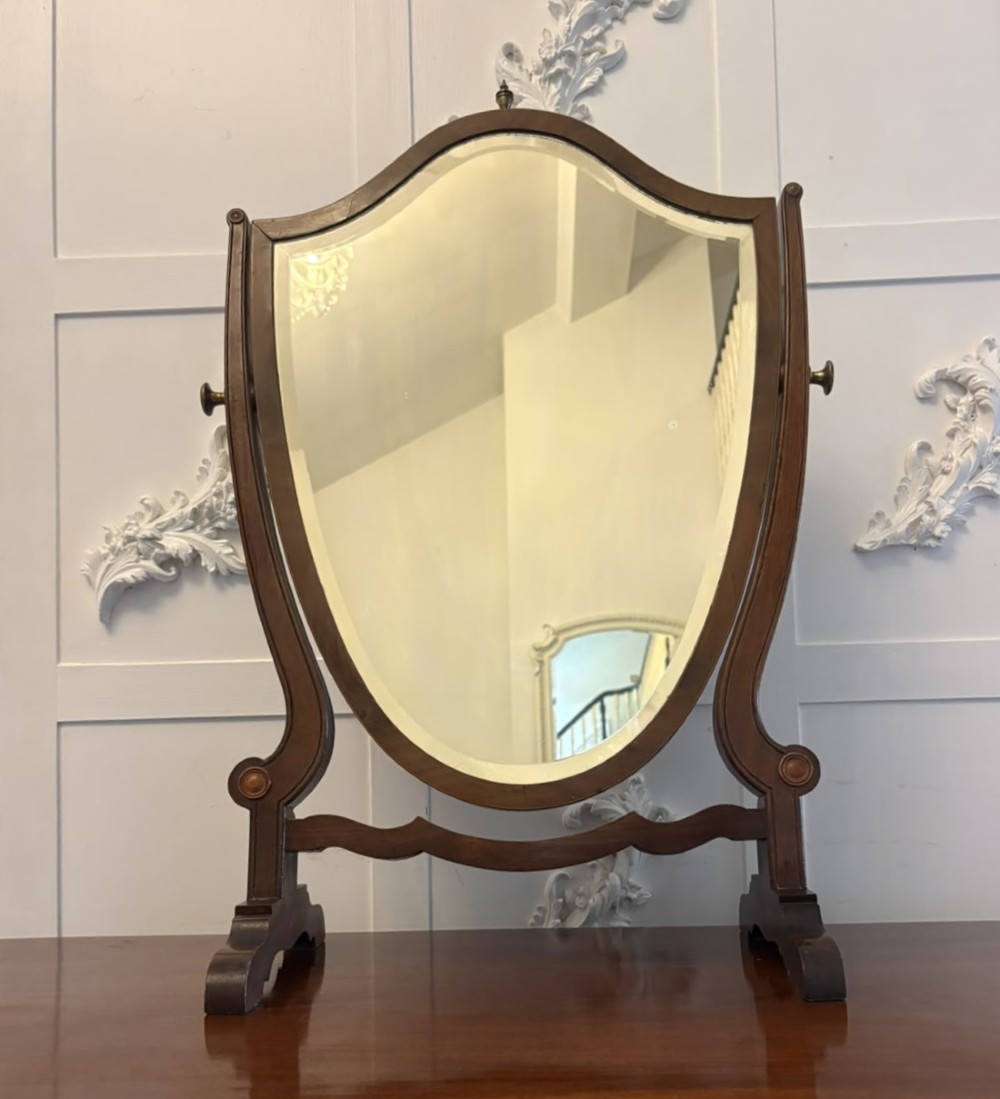 An Edwardian mahogany toilet mirror featuring a shield-shaped frame with bevelled glass mounted on a swivelling stand with brass knobs.