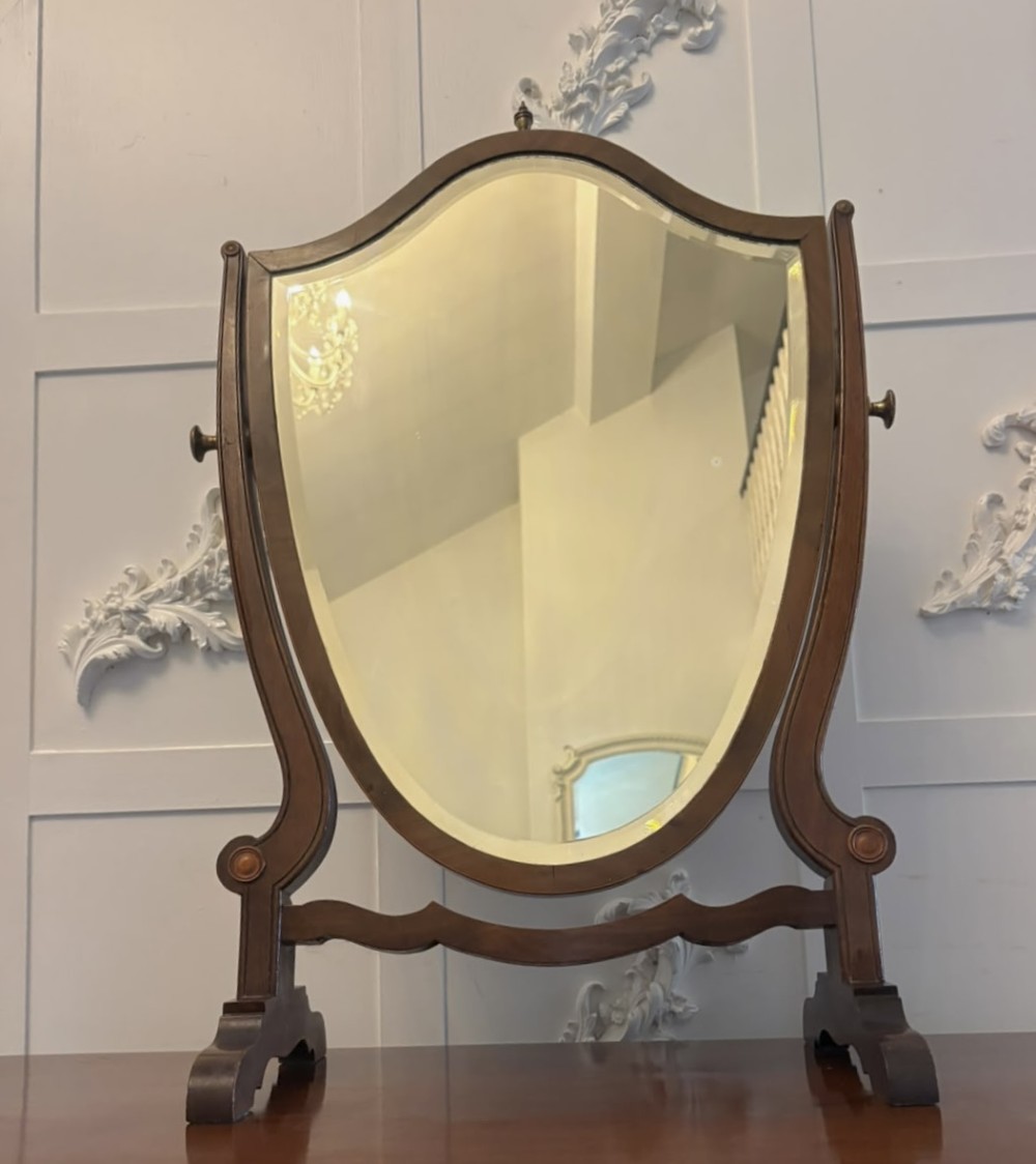An Edwardian mahogany toilet mirror featuring a shield-shaped frame with bevelled glass mounted on a swivelling stand with brass knobs. View VI