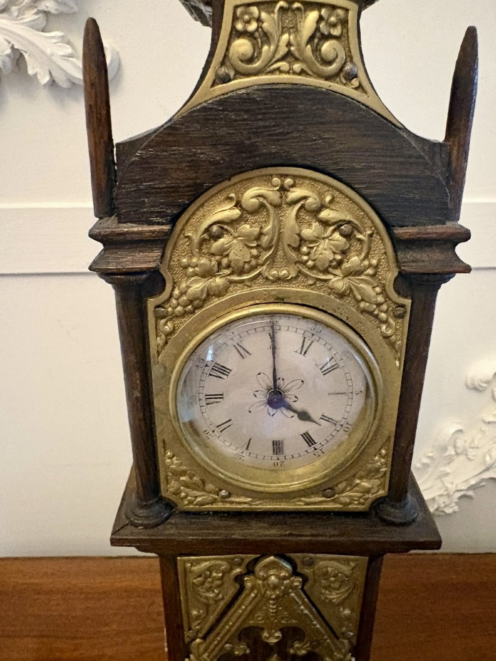 A dark oak miniature grandfather table clock with ornate embossed brass panels and a white dial featuring Roman numerals. View IV