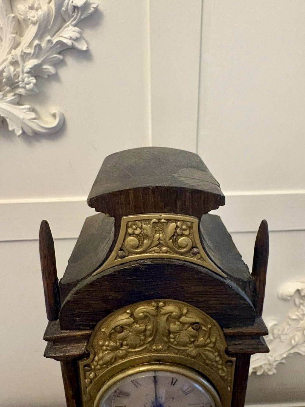 A dark oak miniature grandfather table clock with ornate embossed brass panels and a white dial featuring Roman numerals. View VIII