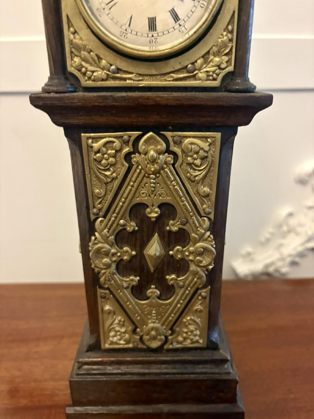 A dark oak miniature grandfather table clock with ornate embossed brass panels and a white dial featuring Roman numerals. View XII