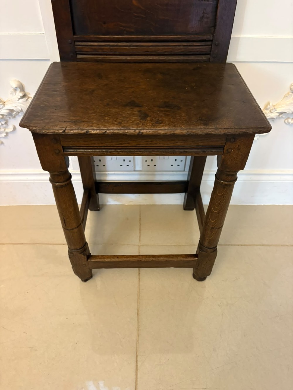A dark oak 17th-century correction chair featuring a tall panelled backrest, carved crest rail with small wooden finials, solid wooden seat, turned front legs, and plain stretchers. View V