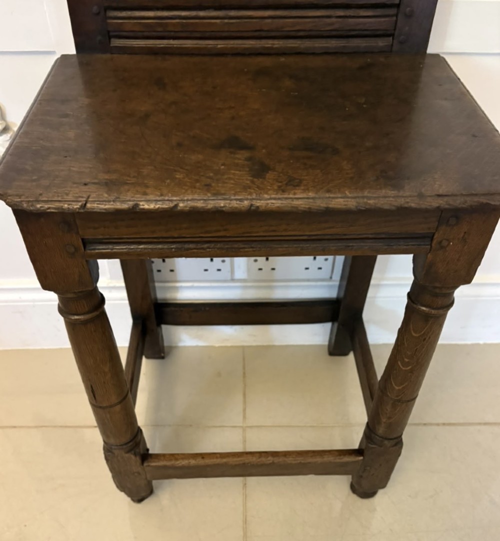 A dark oak 17th-century correction chair featuring a tall panelled backrest, carved crest rail with small wooden finials, solid wooden seat, turned front legs, and plain stretchers. View VI
