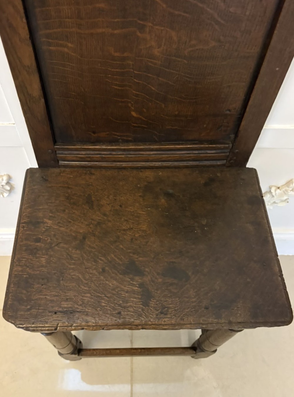 A dark oak 17th-century correction chair featuring a tall panelled backrest, carved crest rail with small wooden finials, solid wooden seat, turned front legs, and plain stretchers. View VII