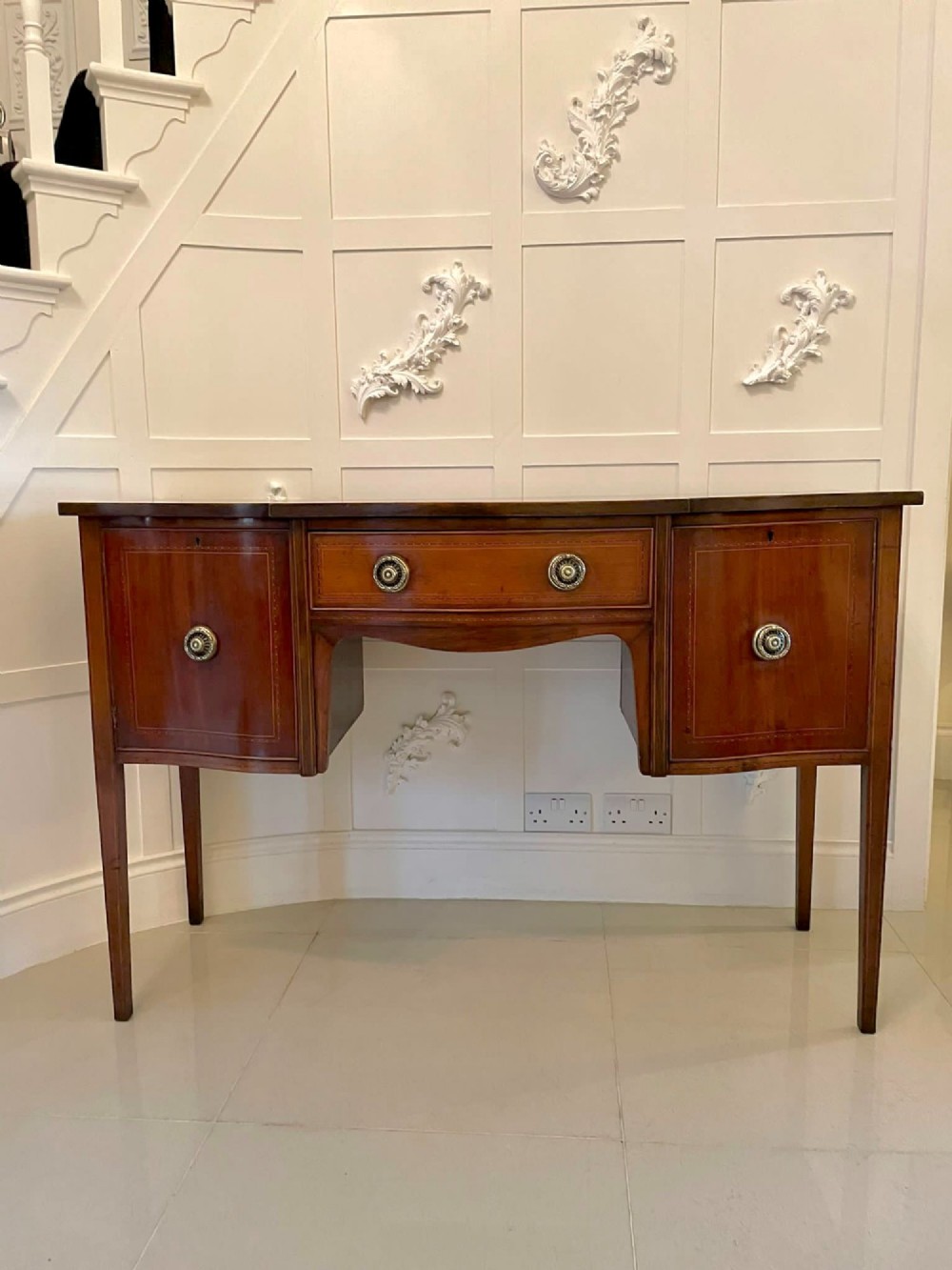 An Edwardian inlaid mahogany serpentine sideboard featuring a central drawer, flanking cupboard doors, brass circular handles, and tapered legs.