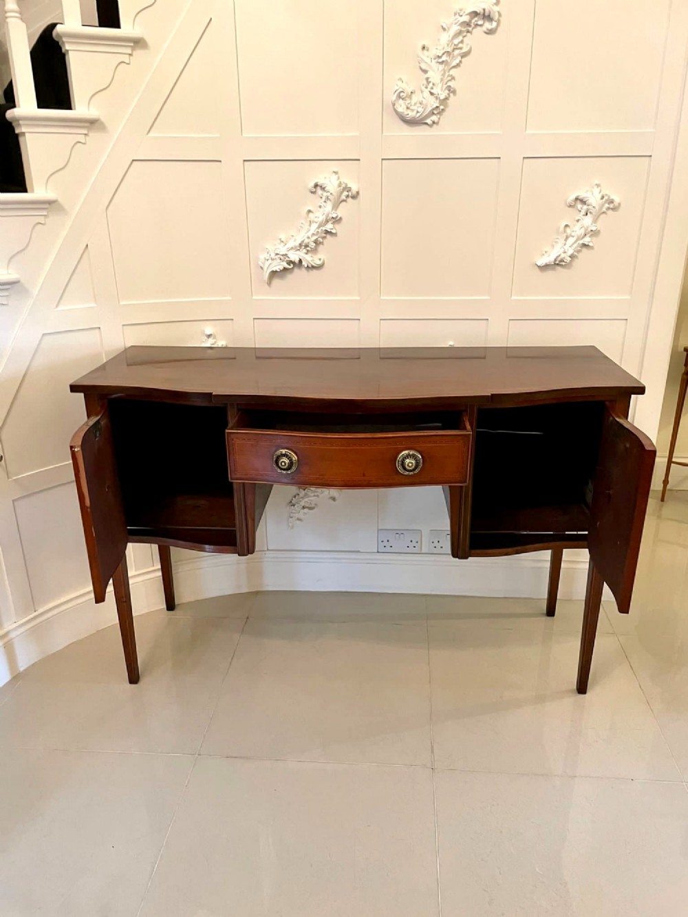 An Edwardian inlaid mahogany serpentine sideboard featuring a central drawer, flanking cupboard doors, brass circular handles, and tapered legs. View V