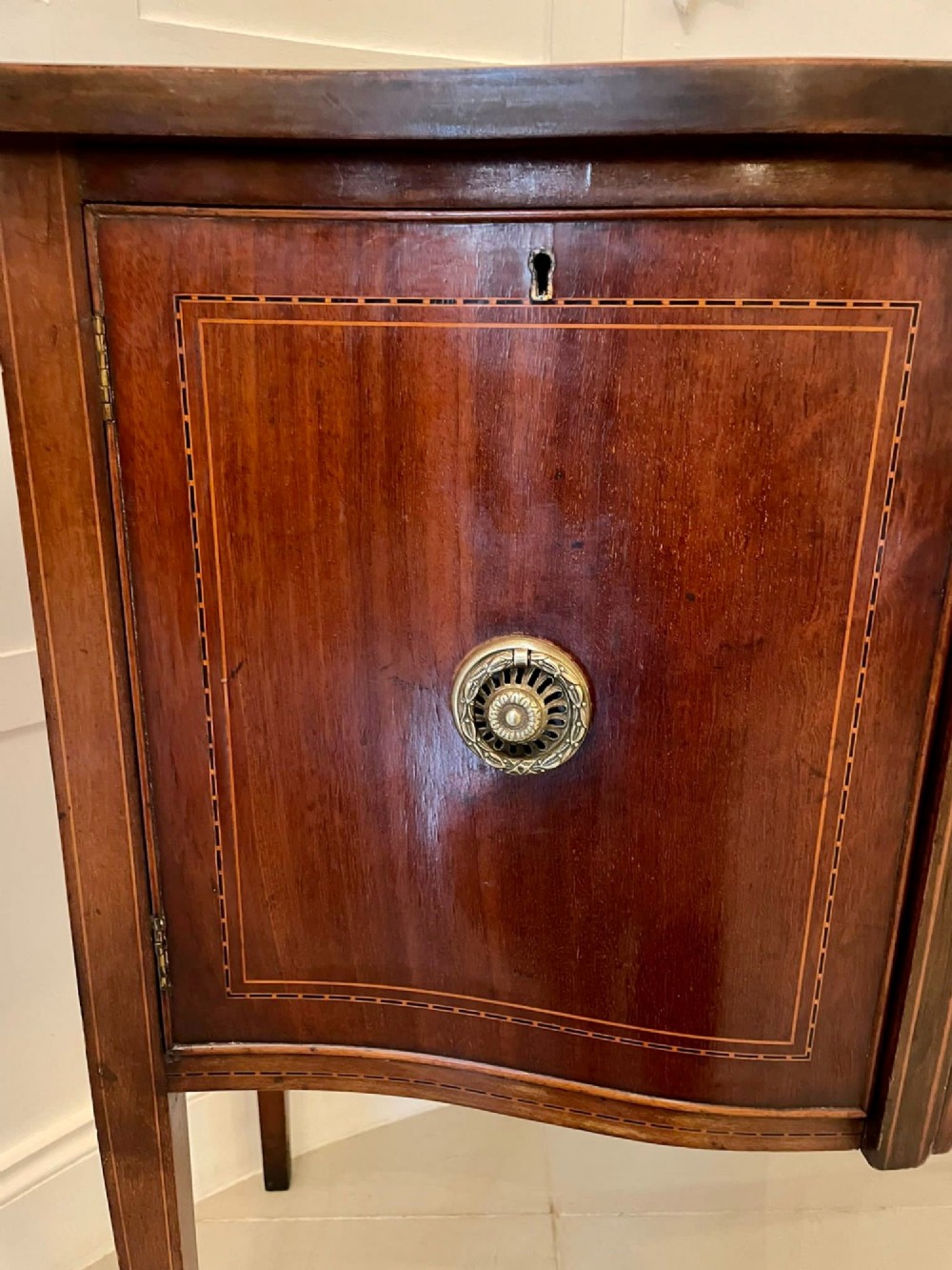 An Edwardian inlaid mahogany serpentine sideboard featuring a central drawer, flanking cupboard doors, brass circular handles, and tapered legs. View XV