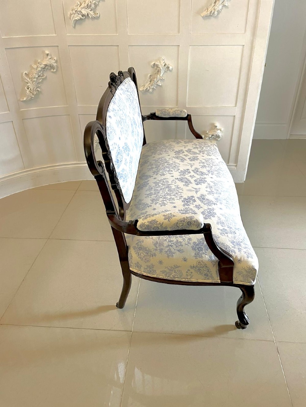 A Victorian carved mahogany settee featuring upholstered blue and white floral pattern seat and backrest, padded armrests, and carved cabriole legs. View VI
