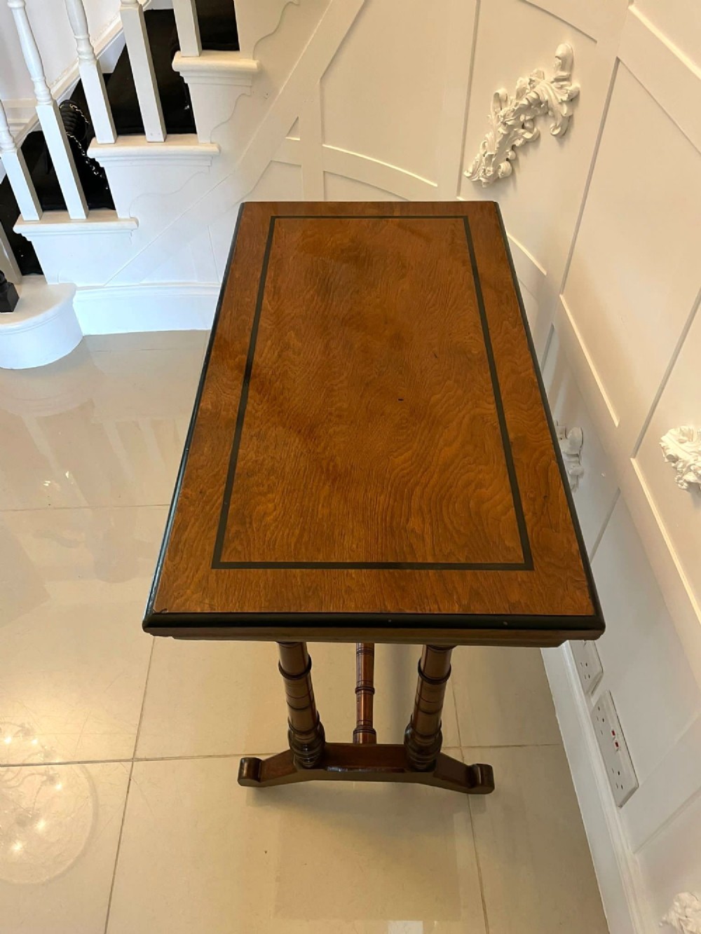 An Arts and Crafts oak side table featuring dark ebony inlay around the rectangular fold-over top, dual turned legs on a stretcher base, and ceramic castors. View X