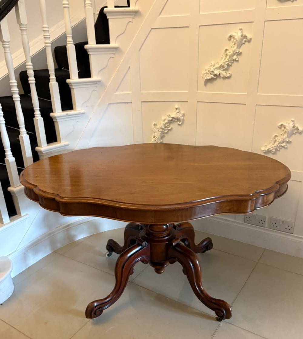 A Victorian figured mahogany tilt-top centre table featuring a shaped serpentine top positioned vertically on a carved central pedestal with four splayed legs. View XII