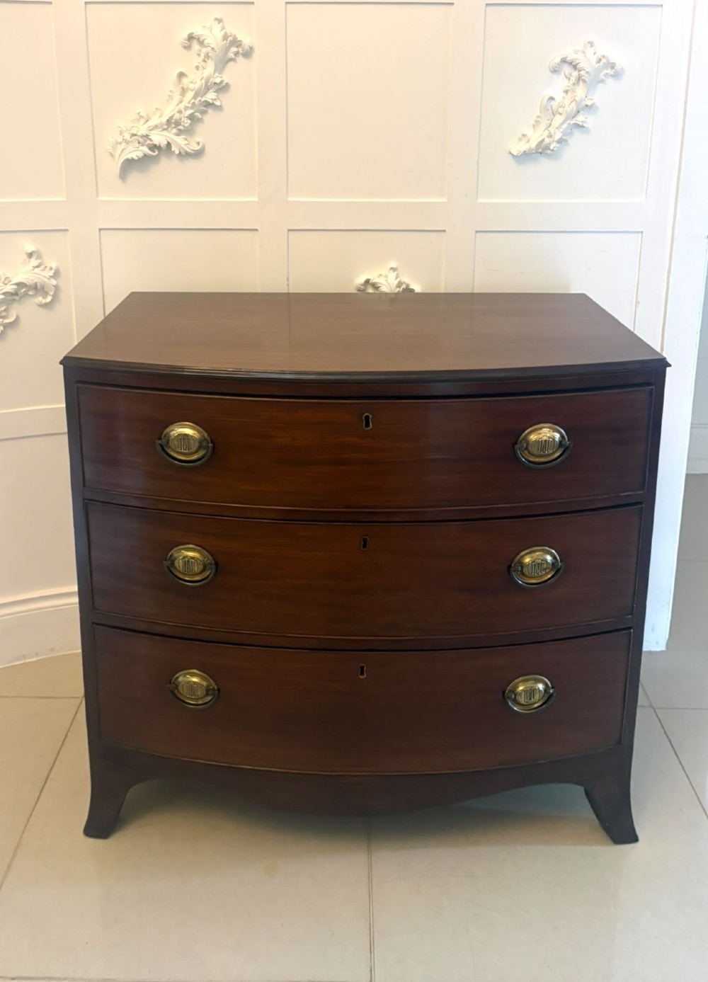 An antique Georgian mahogany bow-front chest featuring three graduating drawers with brass oval handles, supported on splayed feet. View IV