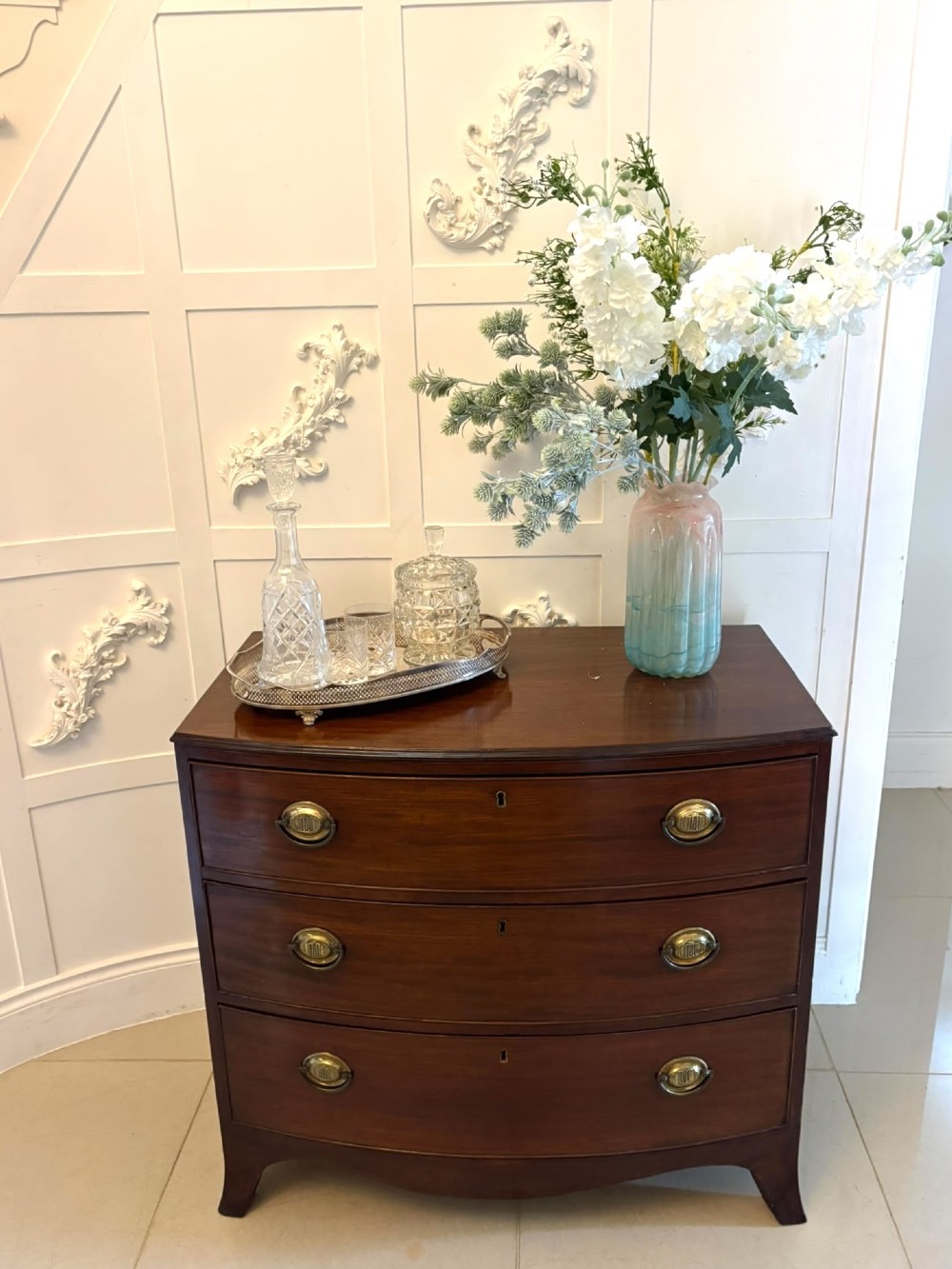 An antique Georgian mahogany bow-front chest featuring three graduating drawers with brass oval handles, supported on splayed feet. View V