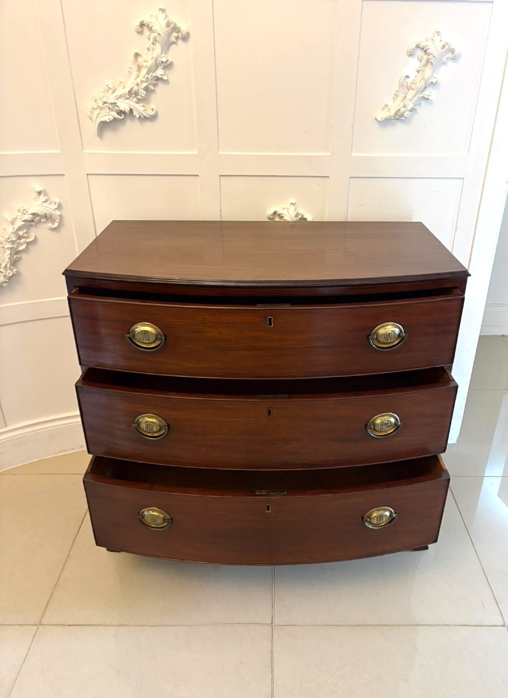 An antique Georgian mahogany bow-front chest featuring three graduating drawers with brass oval handles, supported on splayed feet. View VI
