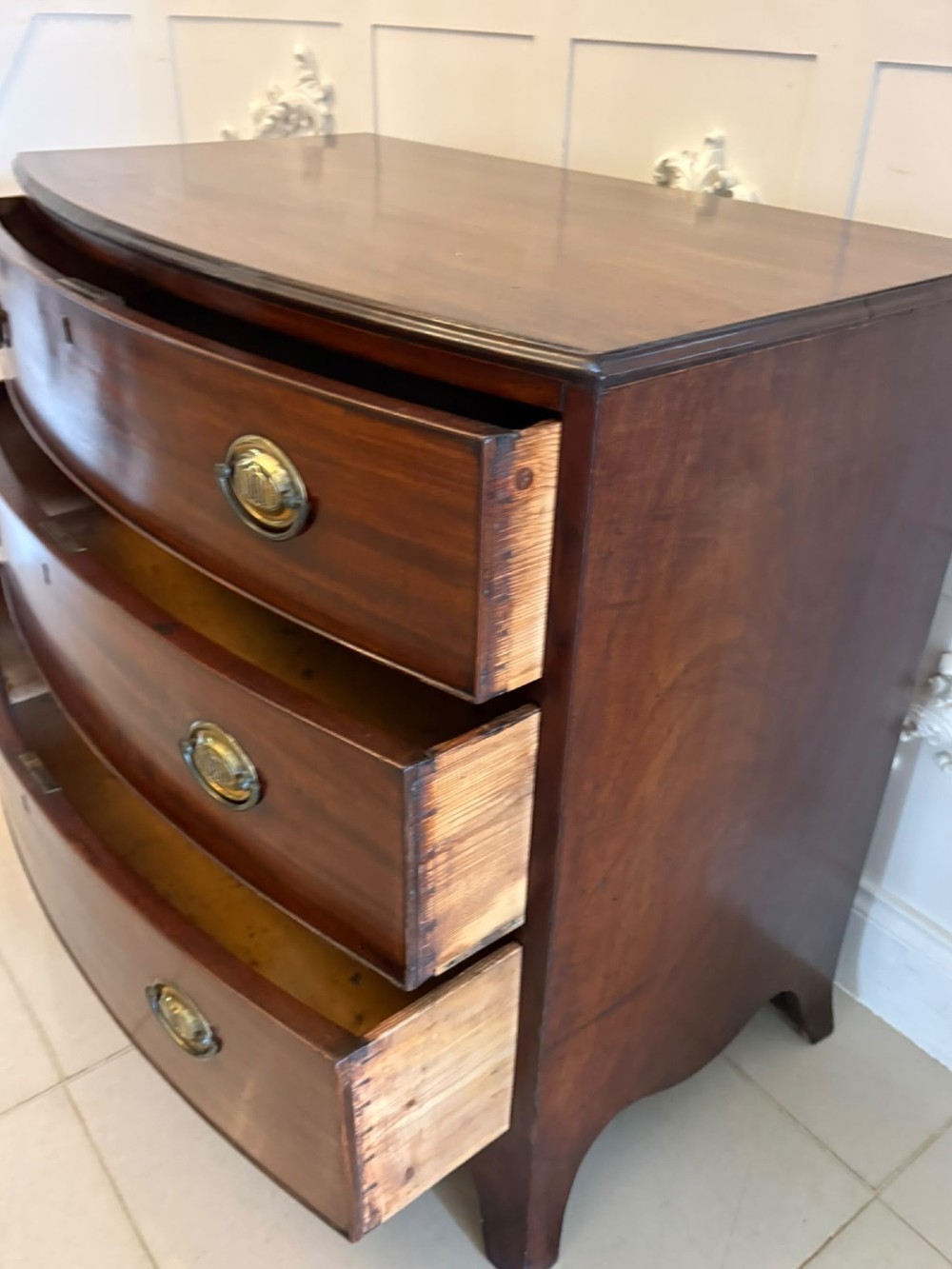 An antique Georgian mahogany bow-front chest featuring three graduating drawers with brass oval handles, supported on splayed feet. View VII
