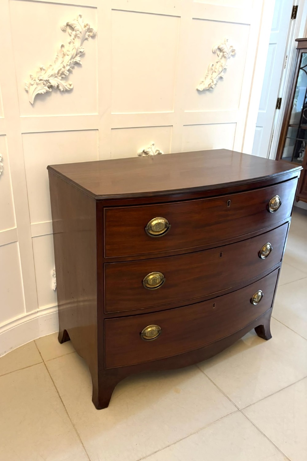 An antique Georgian mahogany bow-front chest featuring three graduating drawers with brass oval handles, supported on splayed feet. View VIII