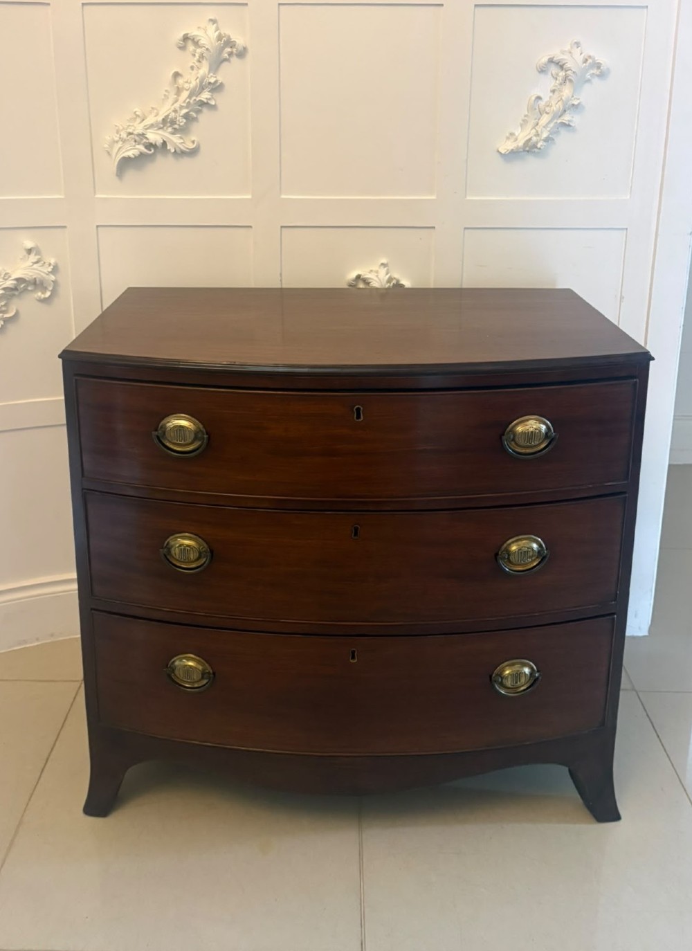 An antique Georgian mahogany bow-front chest featuring three graduating drawers with brass oval handles, supported on splayed feet.