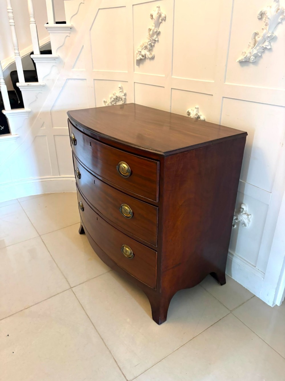 An antique Georgian mahogany bow-front chest featuring three graduating drawers with brass oval handles, supported on splayed feet. View X