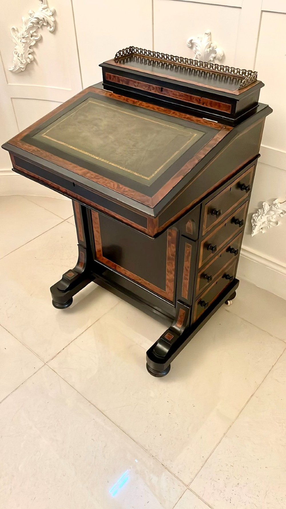A Victorian ebonised davenport desk featuring walnut crossbanding, a sloped green leather writing surface, side drawers, and a top brass gallery rail.