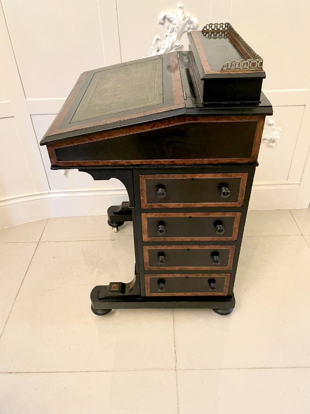 A Victorian ebonised davenport desk featuring walnut crossbanding, a sloped green leather writing surface, side drawers, and a top brass gallery rail. View VII