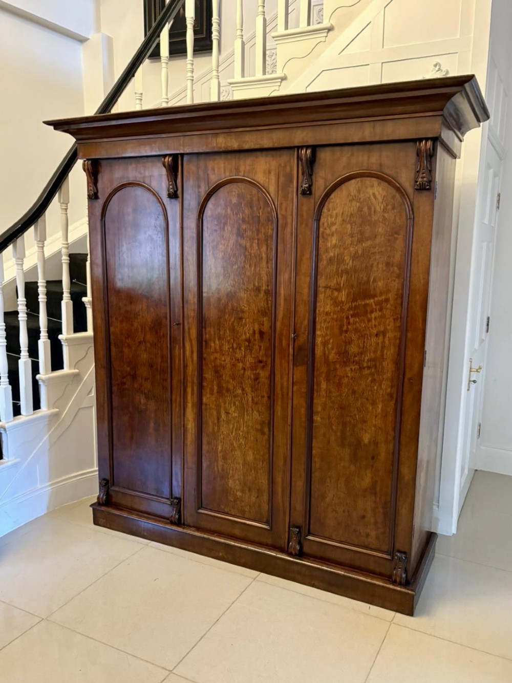 A Victorian three-door wardrobe crafted from figured mahogany with arched door panels, carved top corbels, and a wide moulded cornice. View XIII