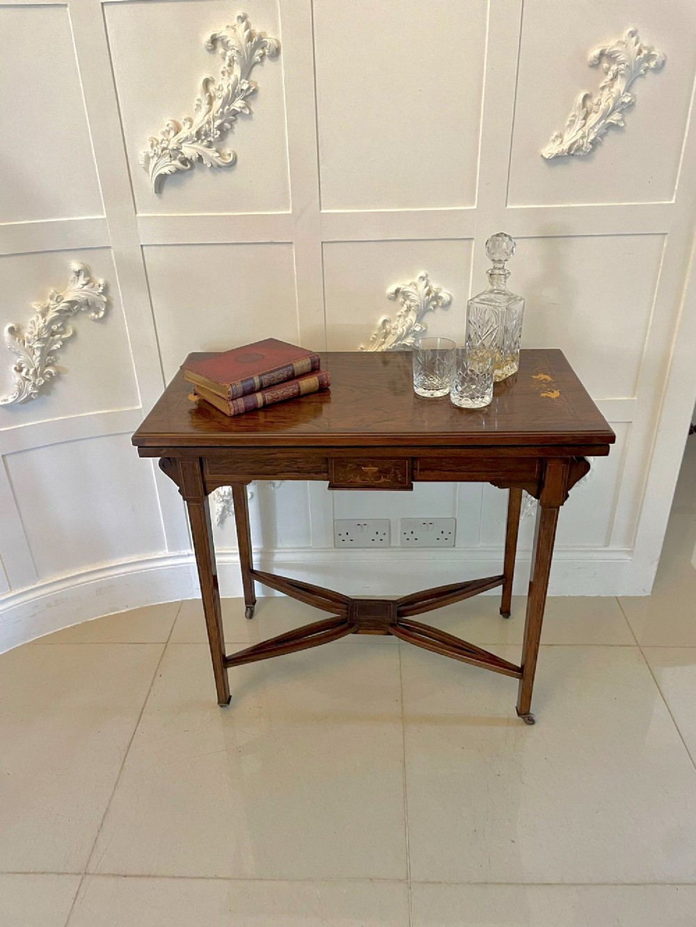 An Edwardian dark rosewood folding card table with floral satinwood corner inlays and an X-frame stretcher resting on brass castors. View II