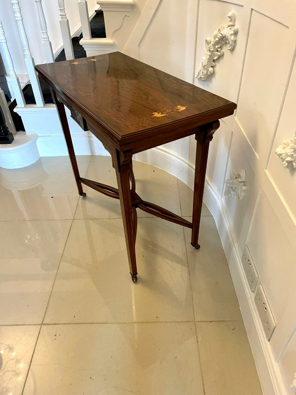 An Edwardian dark rosewood folding card table with floral satinwood corner inlays and an X-frame stretcher resting on brass castors. View VII
