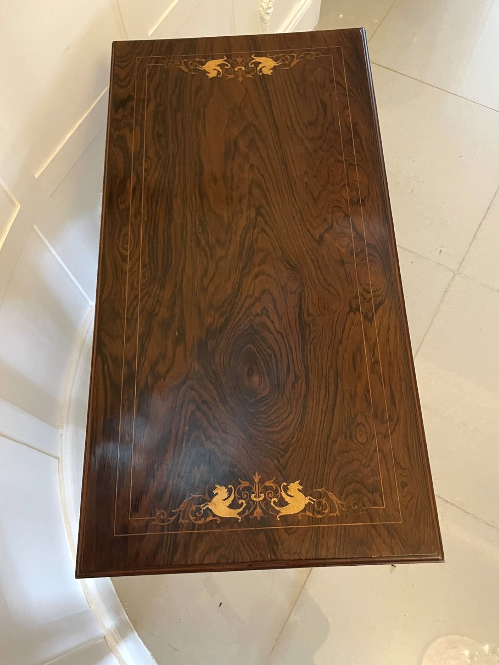 An Edwardian dark rosewood folding card table with floral satinwood corner inlays and an X-frame stretcher resting on brass castors. View X
