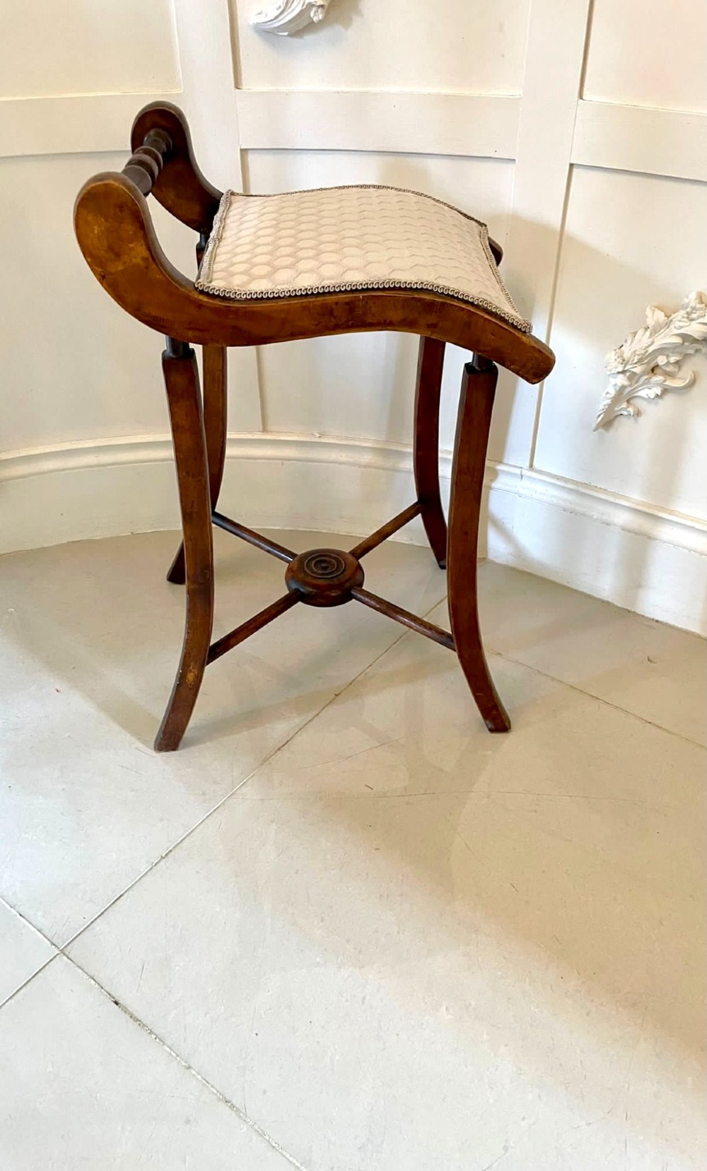 An antique Victorian wooden music chair featuring curved side supports, a turned back rail, splayed legs connected by a central stretcher, and a cream geometric upholstered seat. View VII