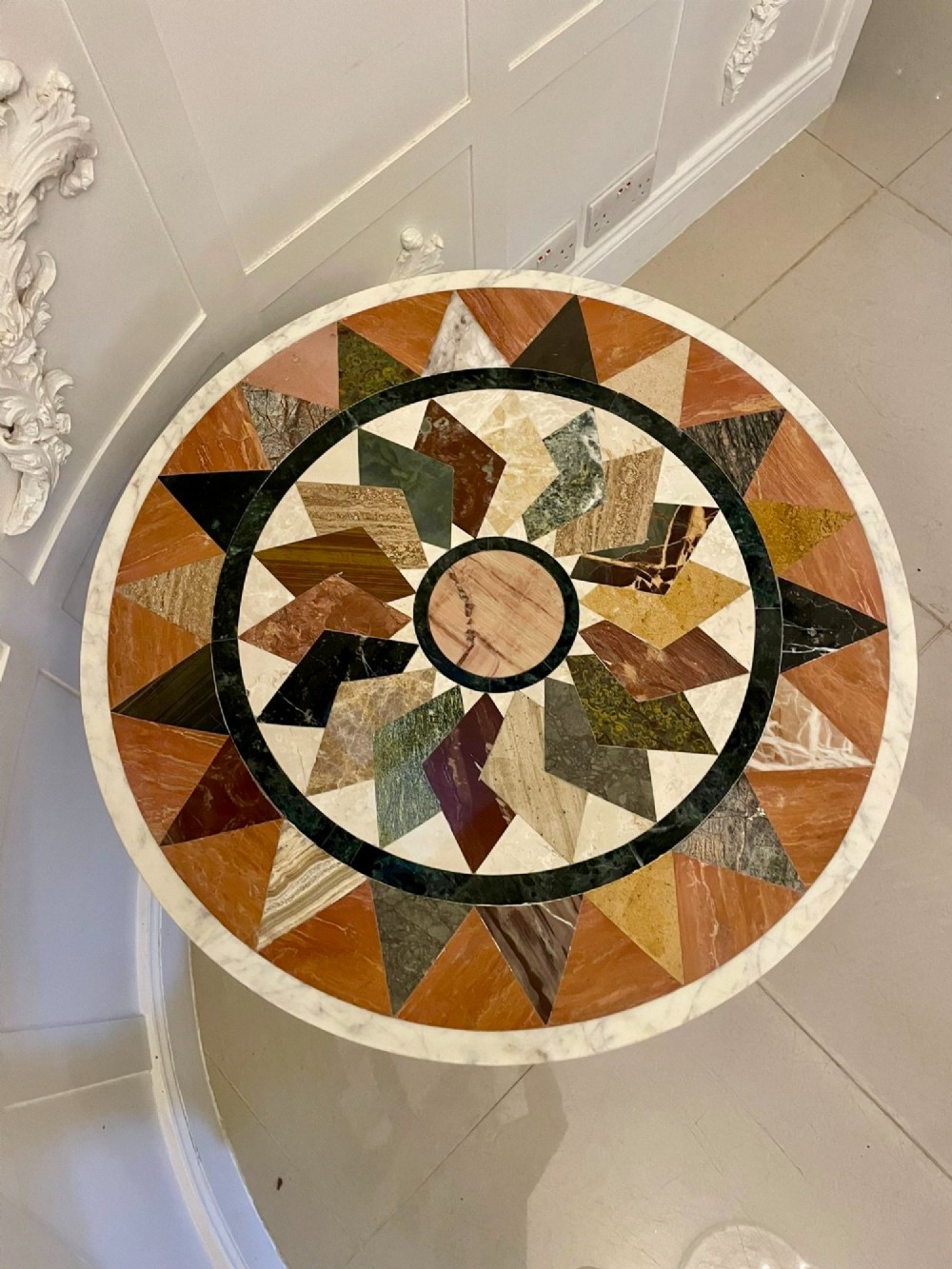 A circular Victorian centre table featuring a geometric specimen marble inlaid top resting on a dark carved mahogany pedestal with a triform base. View IV