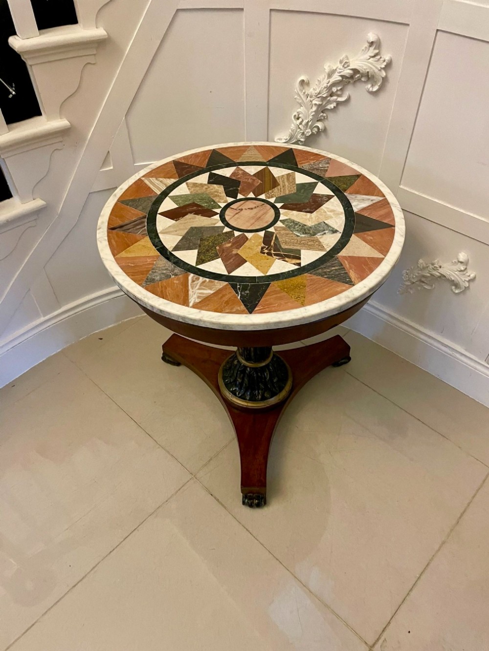 A circular Victorian centre table featuring a geometric specimen marble inlaid top resting on a dark carved mahogany pedestal with a triform base. View VIII