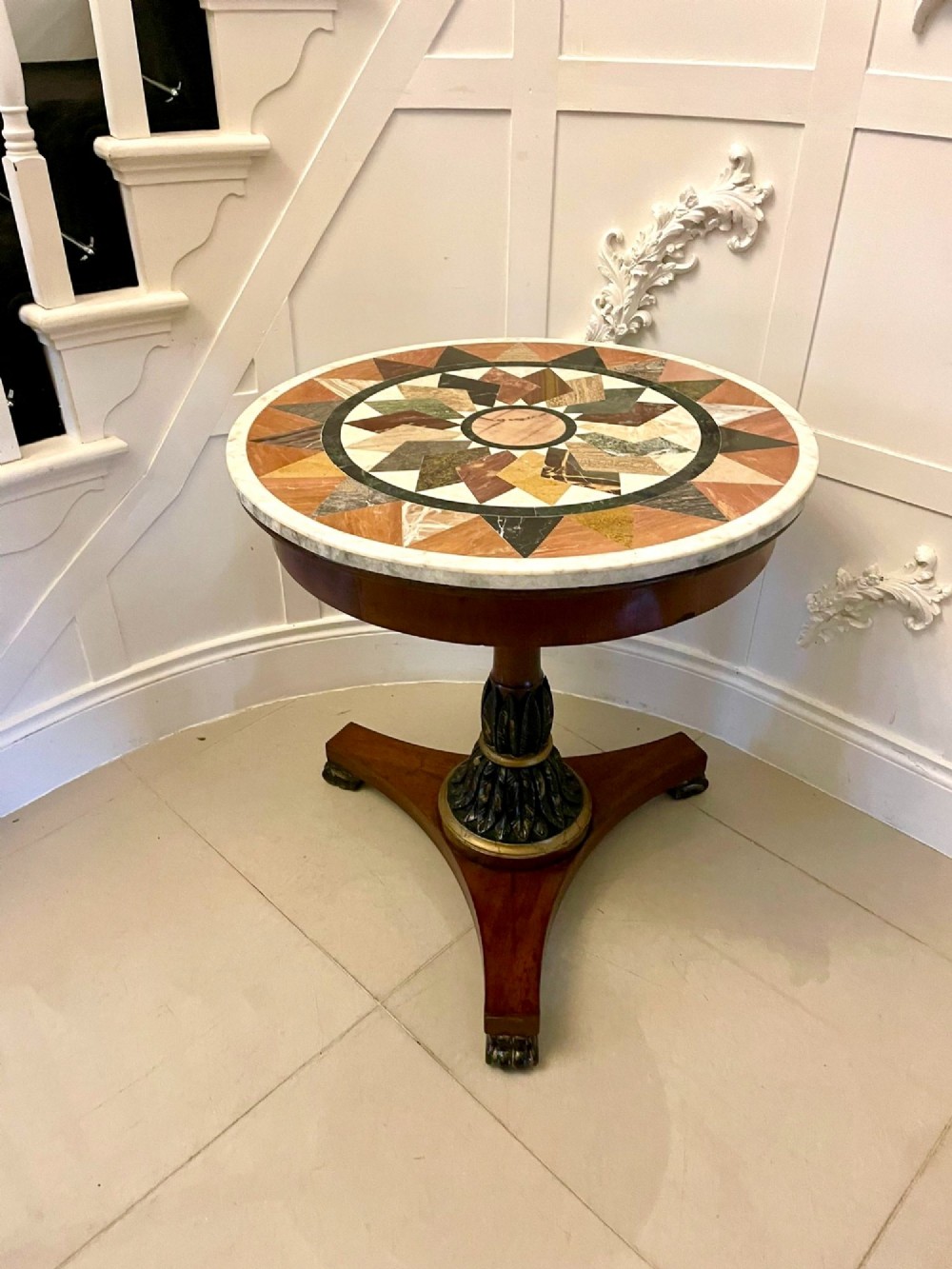 A circular Victorian centre table featuring a geometric specimen marble inlaid top resting on a dark carved mahogany pedestal with a triform base.