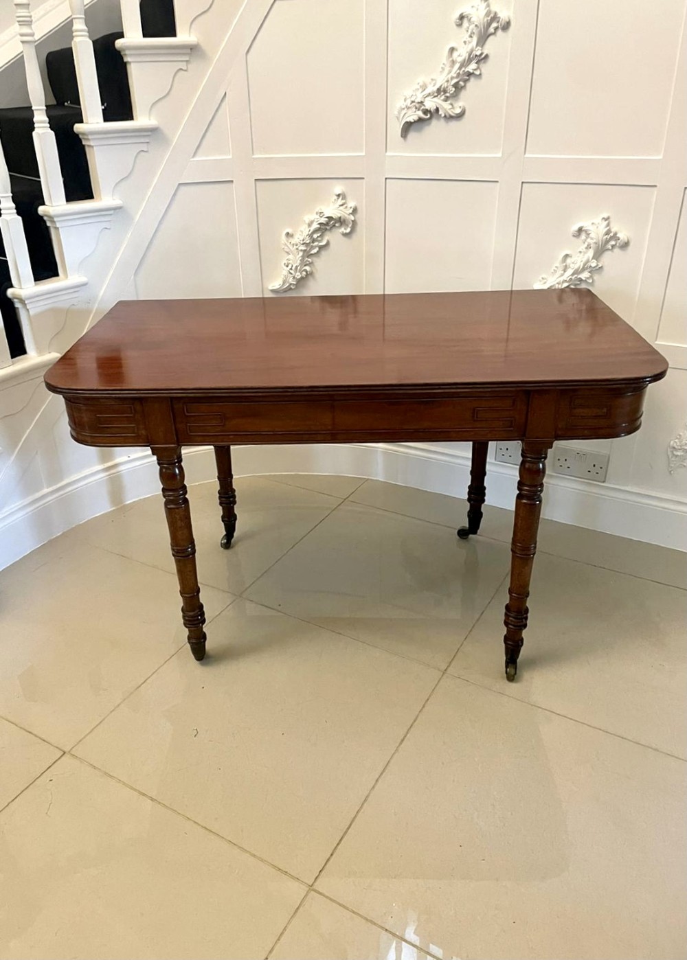 An antique George III mahogany console table featuring a polished rectangular top with rounded corners, a panelled frieze, and turned legs on brass castors.