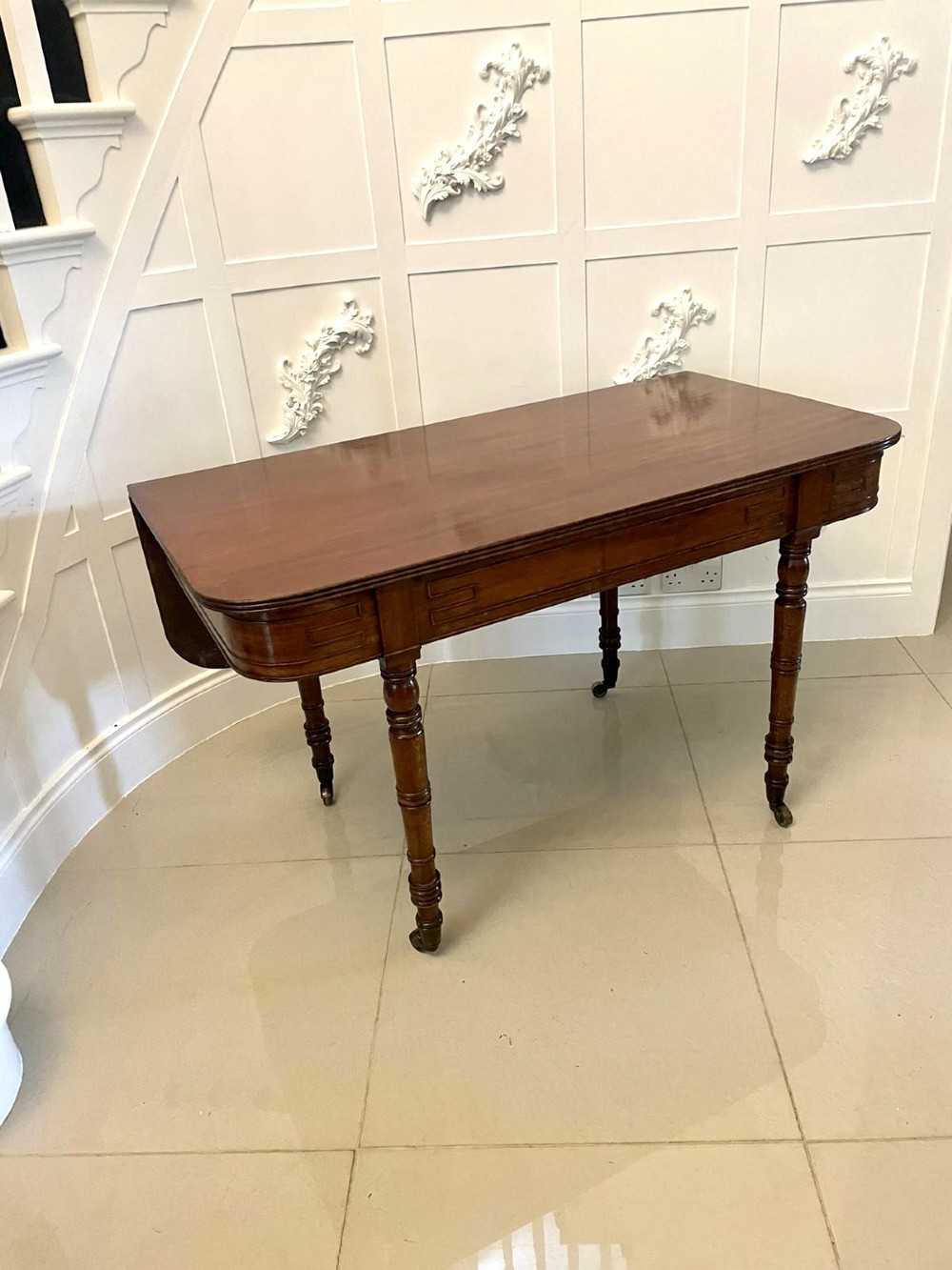 An antique George III mahogany console table featuring a polished rectangular top with rounded corners, a panelled frieze, and turned legs on brass castors. View VII