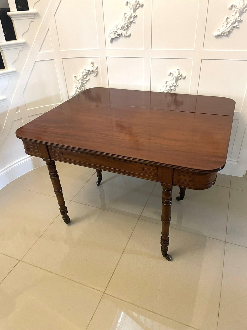 An antique George III mahogany console table featuring a polished rectangular top with rounded corners, a panelled frieze, and turned legs on brass castors. View VIII