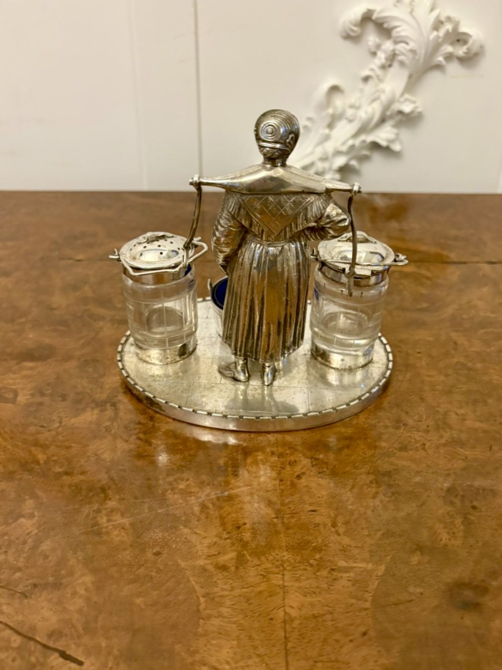An Edwardian silver-plated cruet set featuring a central figural milkmaid holding a yoke with two suspended glass pots and a cobalt blue glass-lined bucket dish on a round tray base. View II