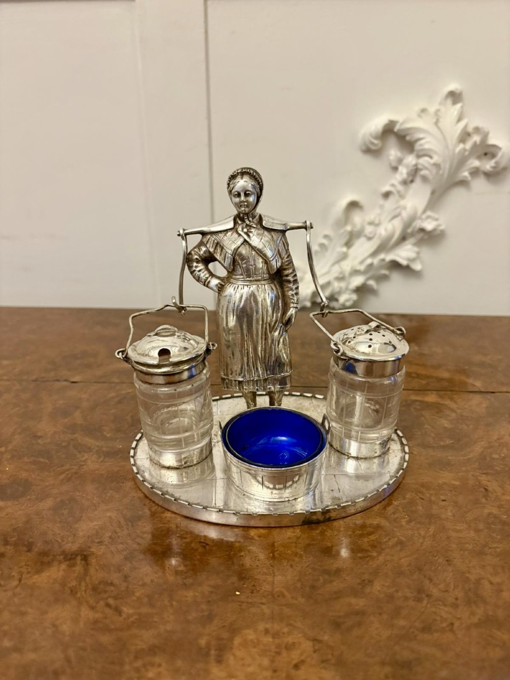 An Edwardian silver-plated cruet set featuring a central figural milkmaid holding a yoke with two suspended glass pots and a cobalt blue glass-lined bucket dish on a round tray base.