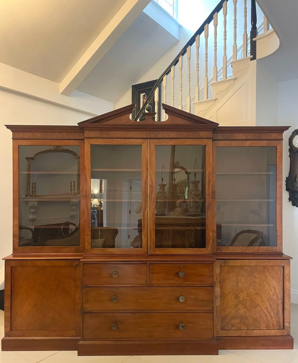 A large George III mahogany breakfront bookcase featuring a swan-neck pediment, glazed upper doors, and a lower section with drawers and cupboards.