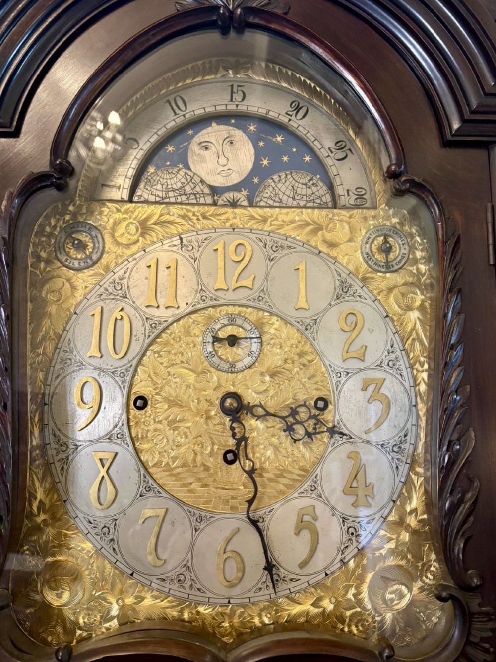 A tall carved dark mahogany longcase clock featuring an ornate pediment, brass dial with moon phase indicator, and a glazed door displaying nine tubular chimes. View VIII