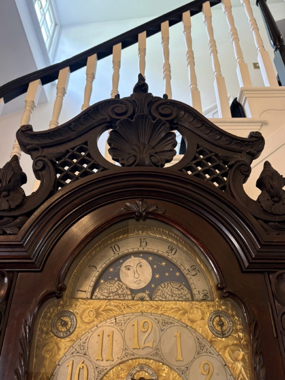 A tall carved dark mahogany longcase clock featuring an ornate pediment, brass dial with moon phase indicator, and a glazed door displaying nine tubular chimes. View IX