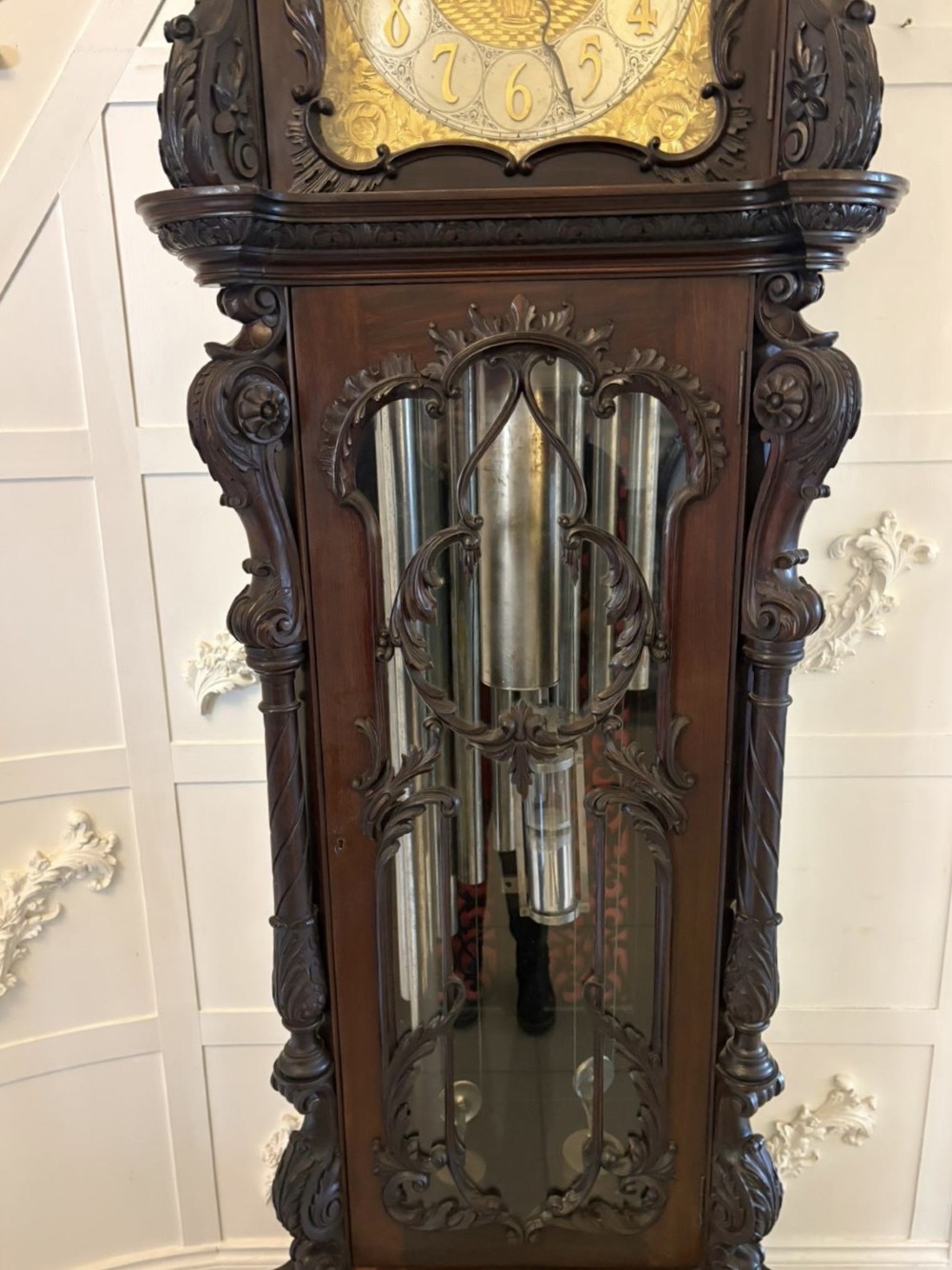 A tall carved dark mahogany longcase clock featuring an ornate pediment, brass dial with moon phase indicator, and a glazed door displaying nine tubular chimes. View XIV