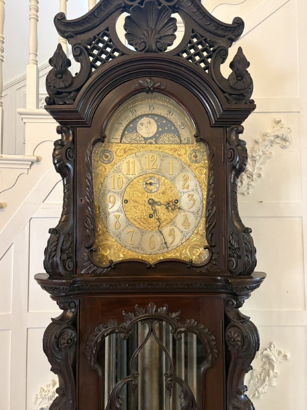 A tall carved dark mahogany longcase clock featuring an ornate pediment, brass dial with moon phase indicator, and a glazed door displaying nine tubular chimes. View XV