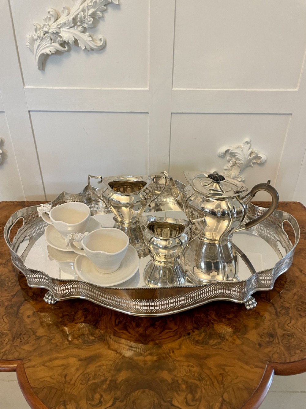 A three-piece Edwardian silver-plated tea set comprising a teapot with a dark wooden handle, a cream jug, and a matching sugar bowl on a polished wood surface. View III