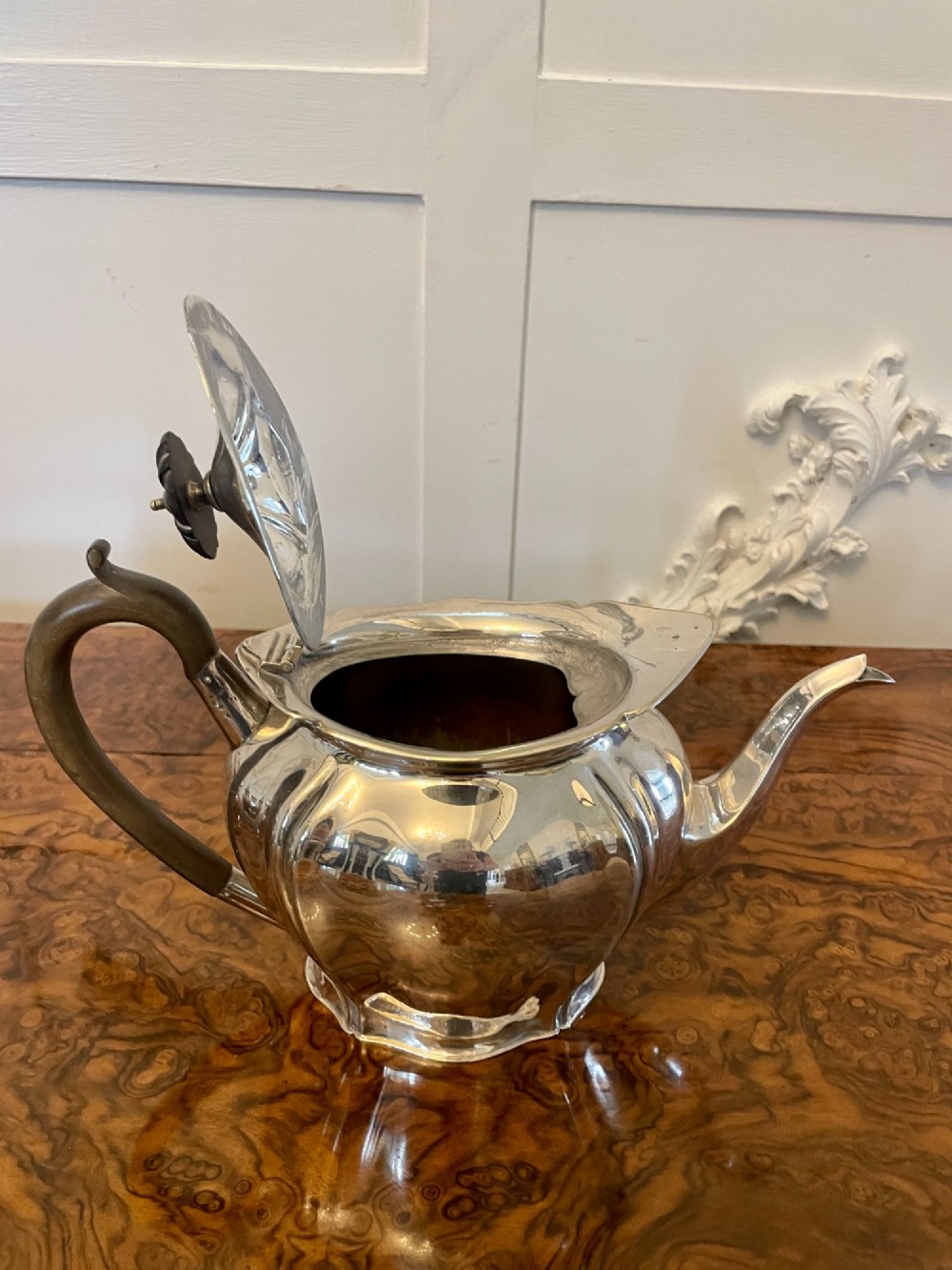 A three-piece Edwardian silver-plated tea set comprising a teapot with a dark wooden handle, a cream jug, and a matching sugar bowl on a polished wood surface. View V