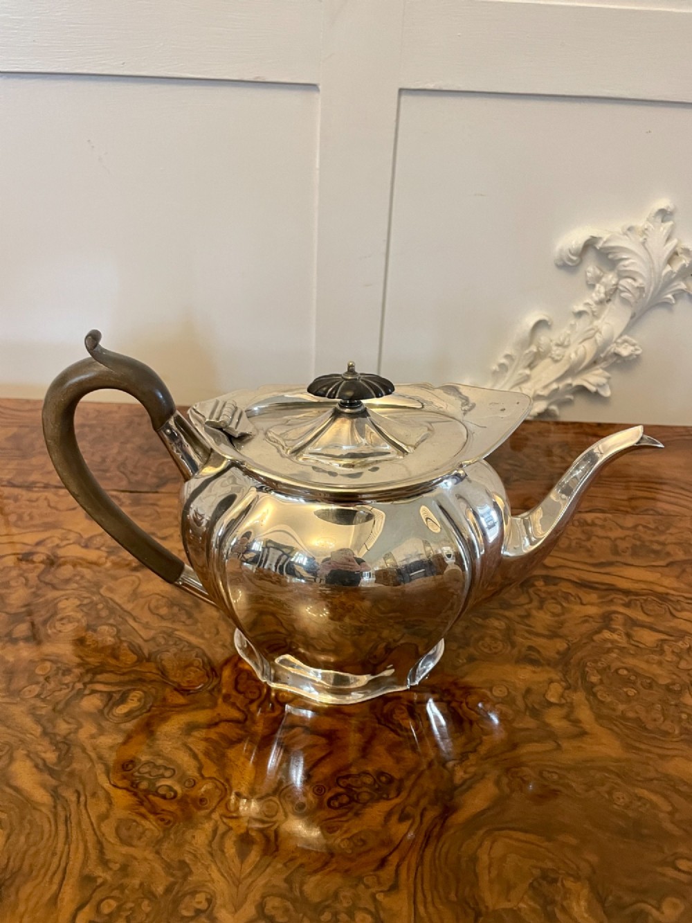 A three-piece Edwardian silver-plated tea set comprising a teapot with a dark wooden handle, a cream jug, and a matching sugar bowl on a polished wood surface. View VI