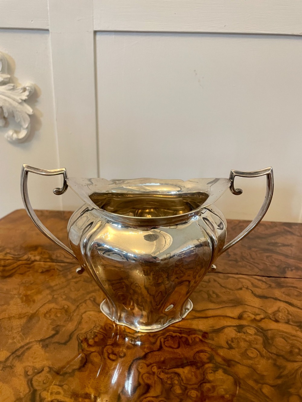 A three-piece Edwardian silver-plated tea set comprising a teapot with a dark wooden handle, a cream jug, and a matching sugar bowl on a polished wood surface. View X