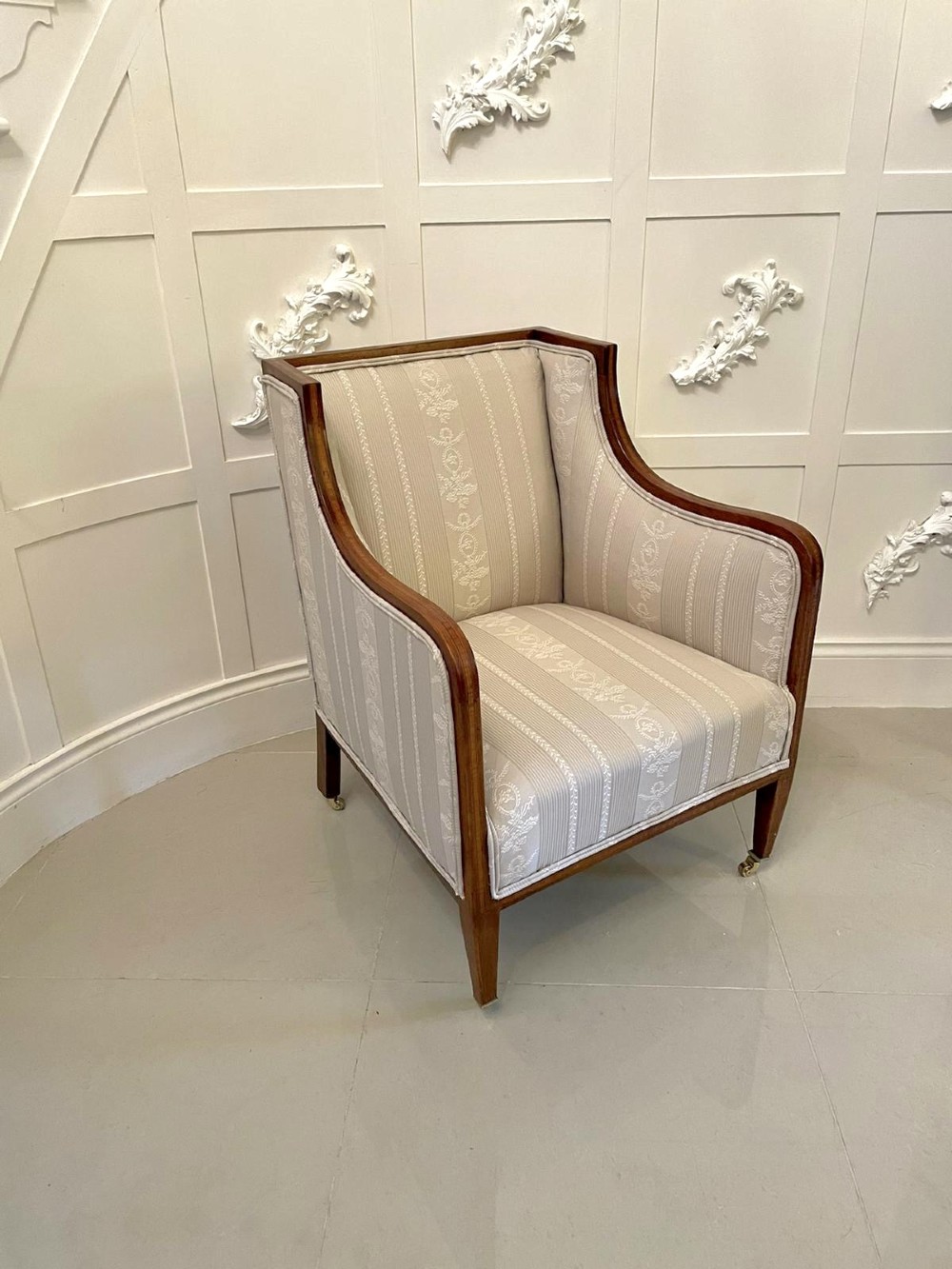 An antique mahogany library armchair upholstered in cream striped fabric, featuring fine timber inlay on the sweeping frame and tapered legs on brass castors.